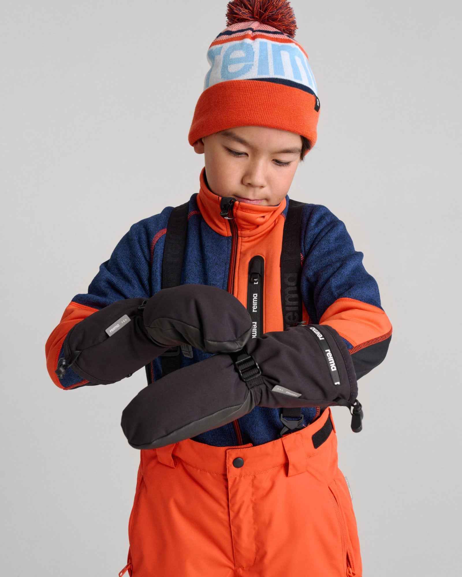 Lifestyle portrait of a young boy adjusting his black Reima Snoukka kids' ski mittens on the slopes. The image demonstrates how the extra-long cuffs easily layer over a winter jacket to completely block out wind and snow.