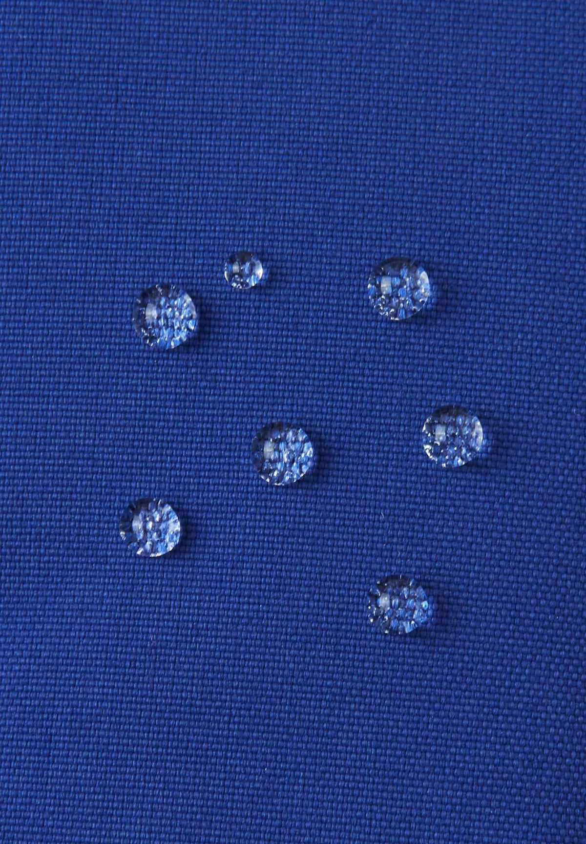 Macro close-up of water droplets actively beading and rolling off the twilight blue outer shell of the Reima Snoukka kids' ski mittens, demonstrating the high-performance waterproof and breathable outer membrane.
