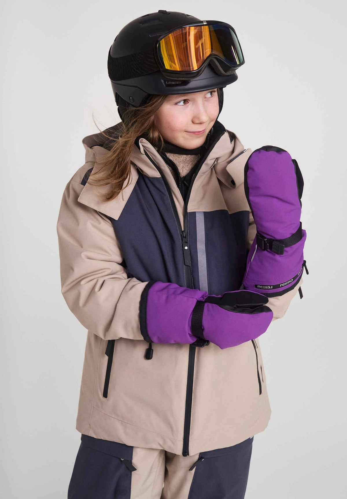 Lifestyle portrait of a young girl ready for winter sports in an earthy beige ski jacket, black alpine helmet, and goggles, wearing thick plum purple Reima Snoukka kids' ski mittens. The image showcases the mittens' long protective cuffs and adjustable wrist straps designed to seal tightly over jacket sleeves and lock out deep snow.