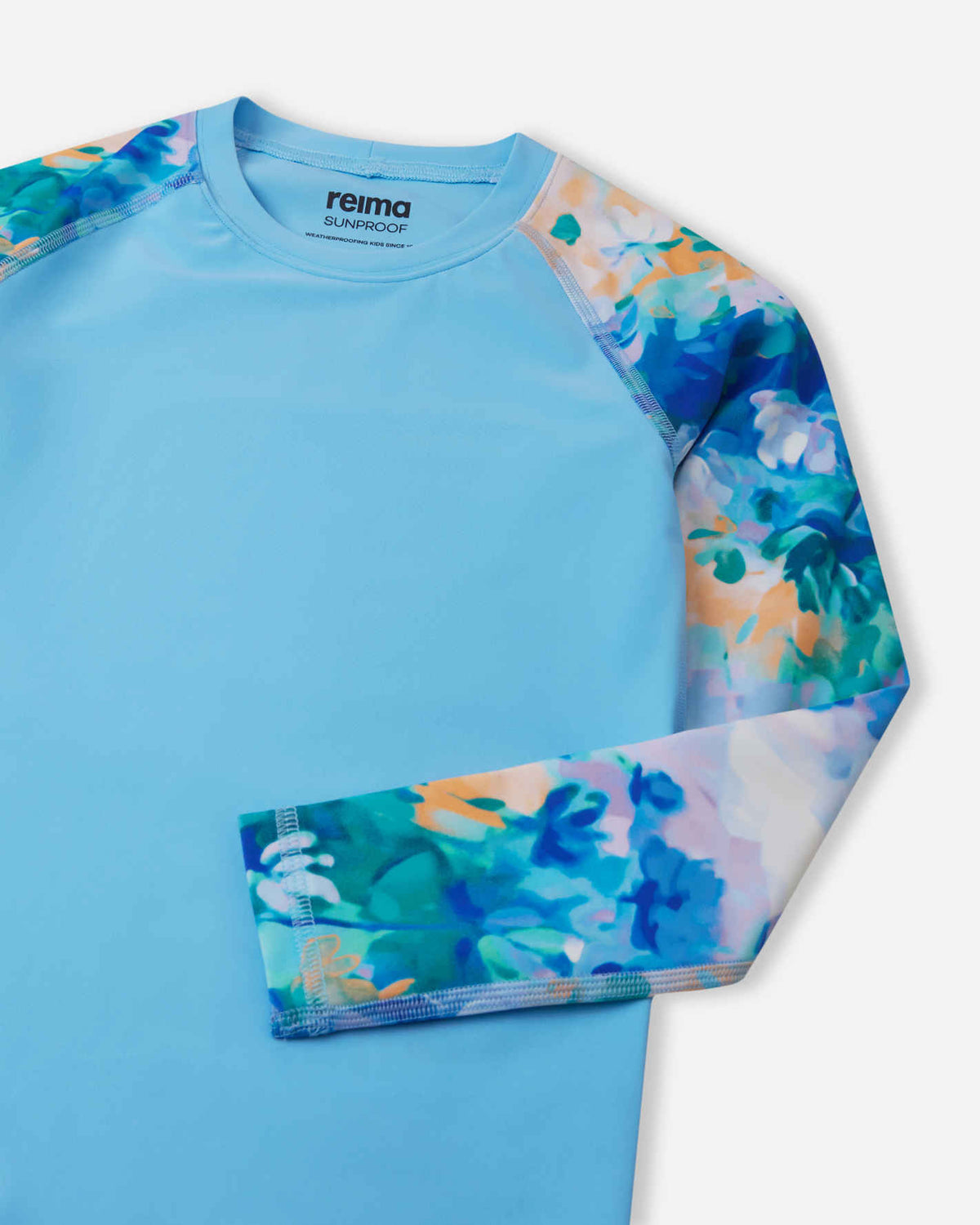 Close-up detail of the Reima Sukeltaja kids' sunproof rash guard in fresh blue, highlighting the vibrant watercolor floral sleeve, smooth flatlock stitching to prevent chafing, and a protective stretch crew neckline.
