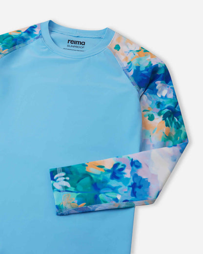 Close-up detail of the Reima Sukeltaja kids' sunproof rash guard in fresh blue, highlighting the vibrant watercolor floral sleeve, smooth flatlock stitching to prevent chafing, and a protective stretch crew neckline.