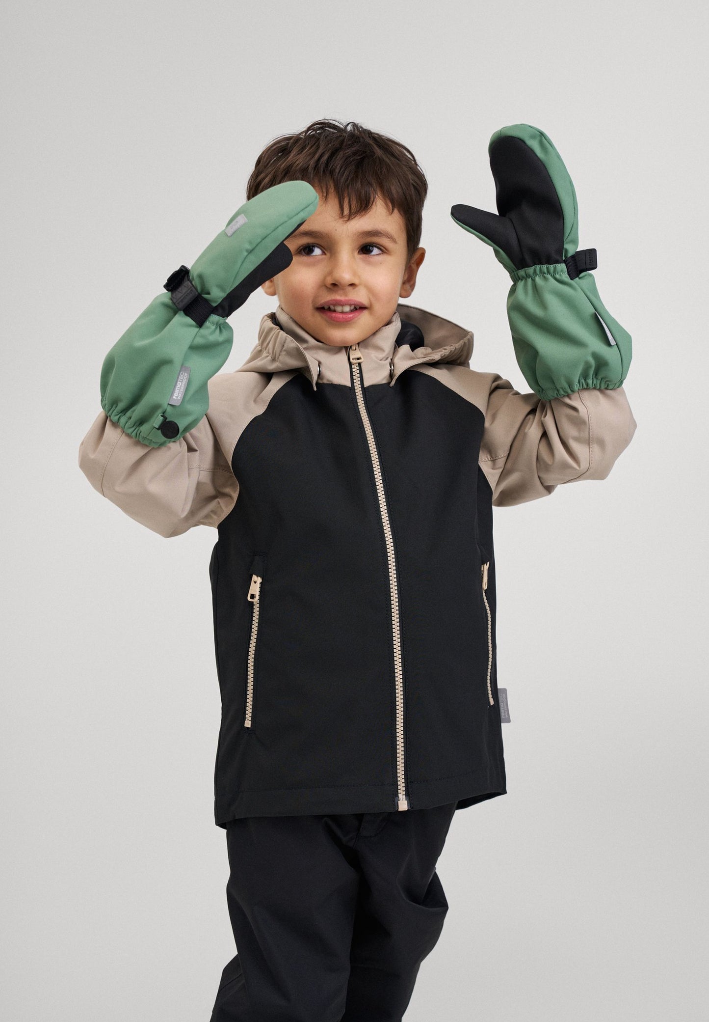 Lifestyle portrait of a young boy smiling and modeling the black and beige Reima Sulkava fall jacket, paired with long green winter mittens, demonstrating a comfortable and warm fit for outdoor play.