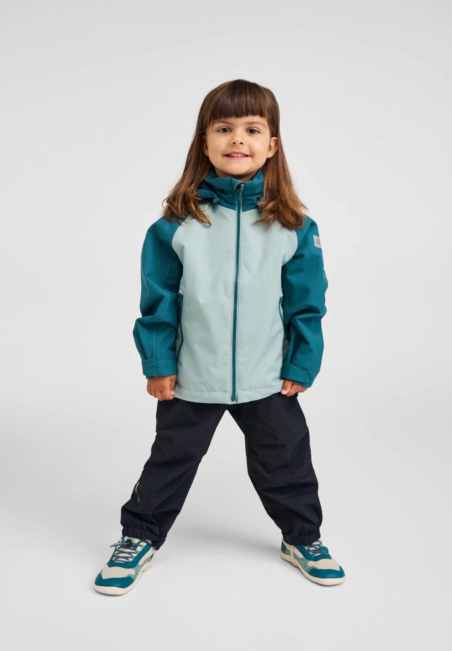 Full-body lifestyle shot of a young girl wearing the color-block stone green Reima Sulkava kids' fall jacket paired with dark outdoor pants and sneakers, perfectly outfitted for active autumn adventures.