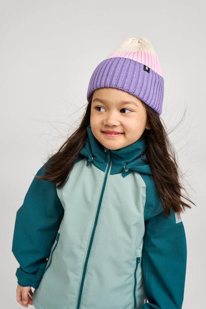 Lifestyle portrait of a young girl smiling and modeling the stone green Reima Sulkava fall jacket, paired with a colorful striped knit beanie, demonstrating a comfortable and stylish fit for crisp autumn weather.