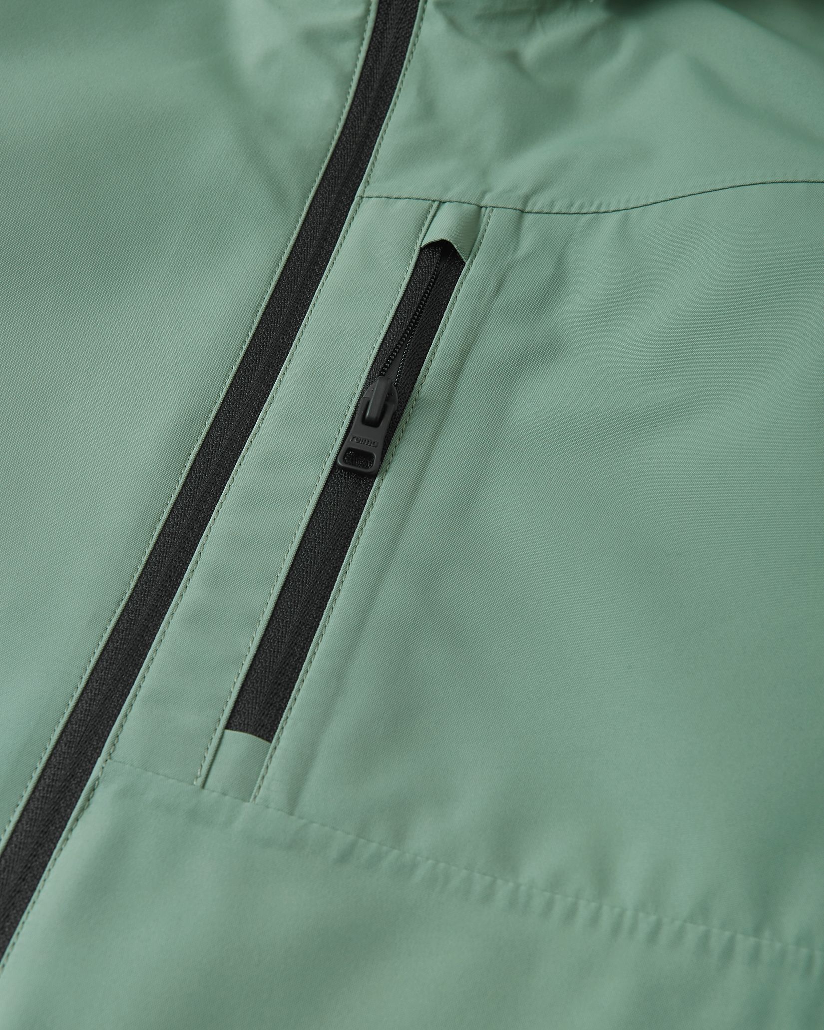 Close-up detail of the Reima Suojala kids' stone green shell jacket, highlighting the secure zippered chest pocket and the smooth, durable windproof fabric