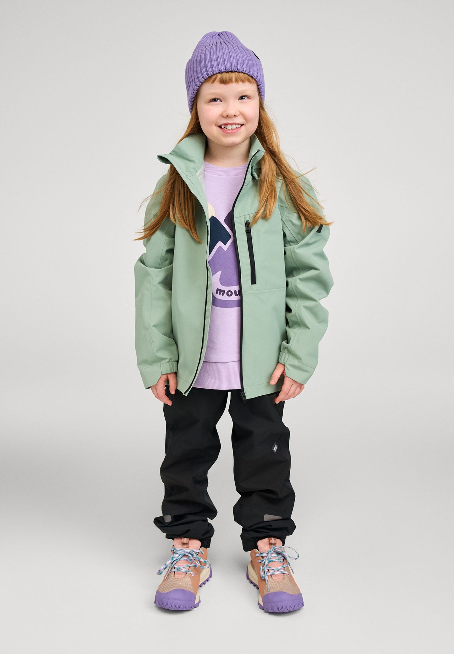 Lifestyle shot of a smiling girl wearing the stone green Reima Suojala kids' waterproof shell jacket paired with black outdoor pants and a purple beanie, demonstrating its comfortable, active fit.