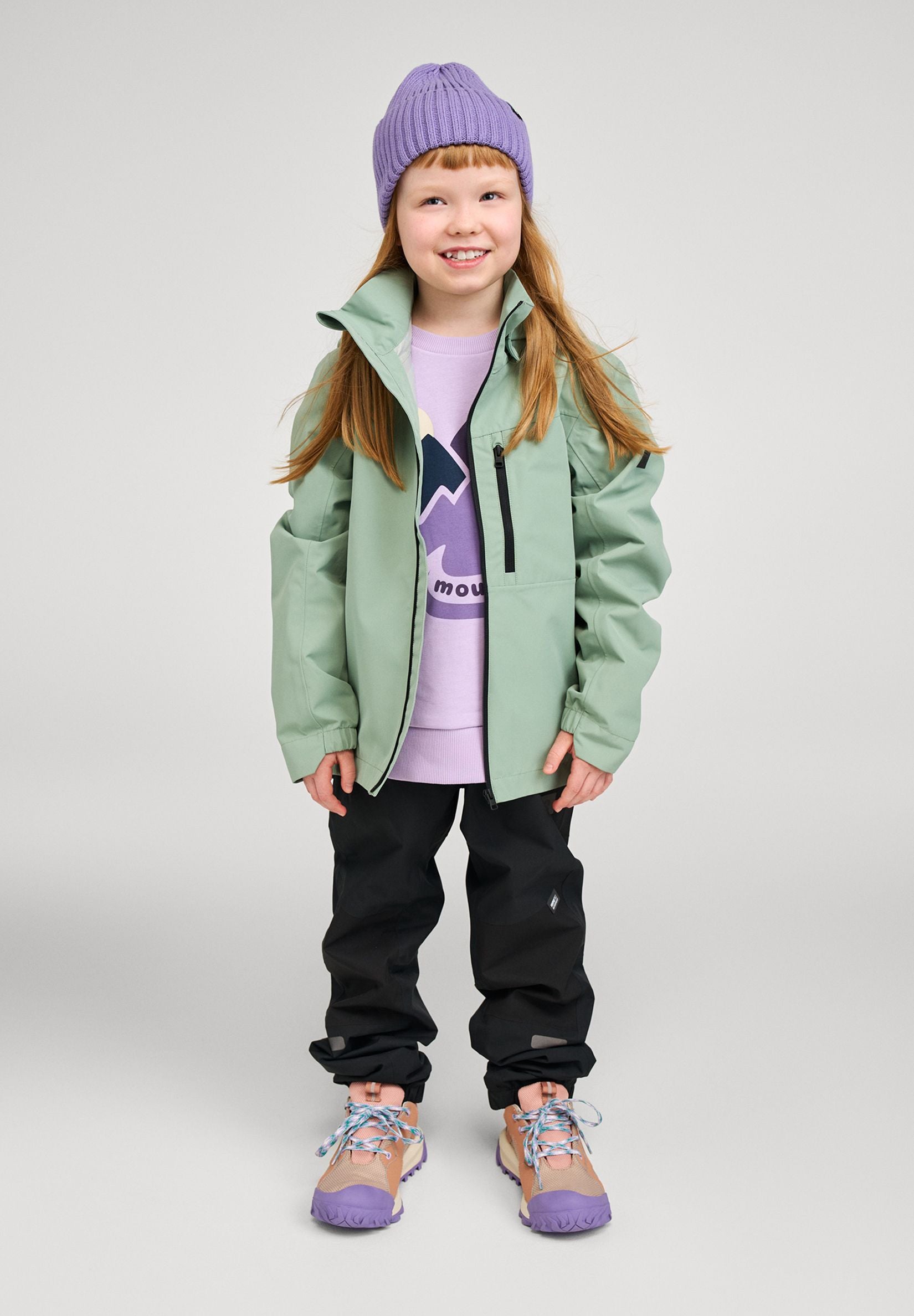 Lifestyle shot of a smiling girl wearing the stone green Reima Suojala kids' waterproof shell jacket paired with black outdoor pants and a purple beanie, demonstrating its comfortable, active fit.