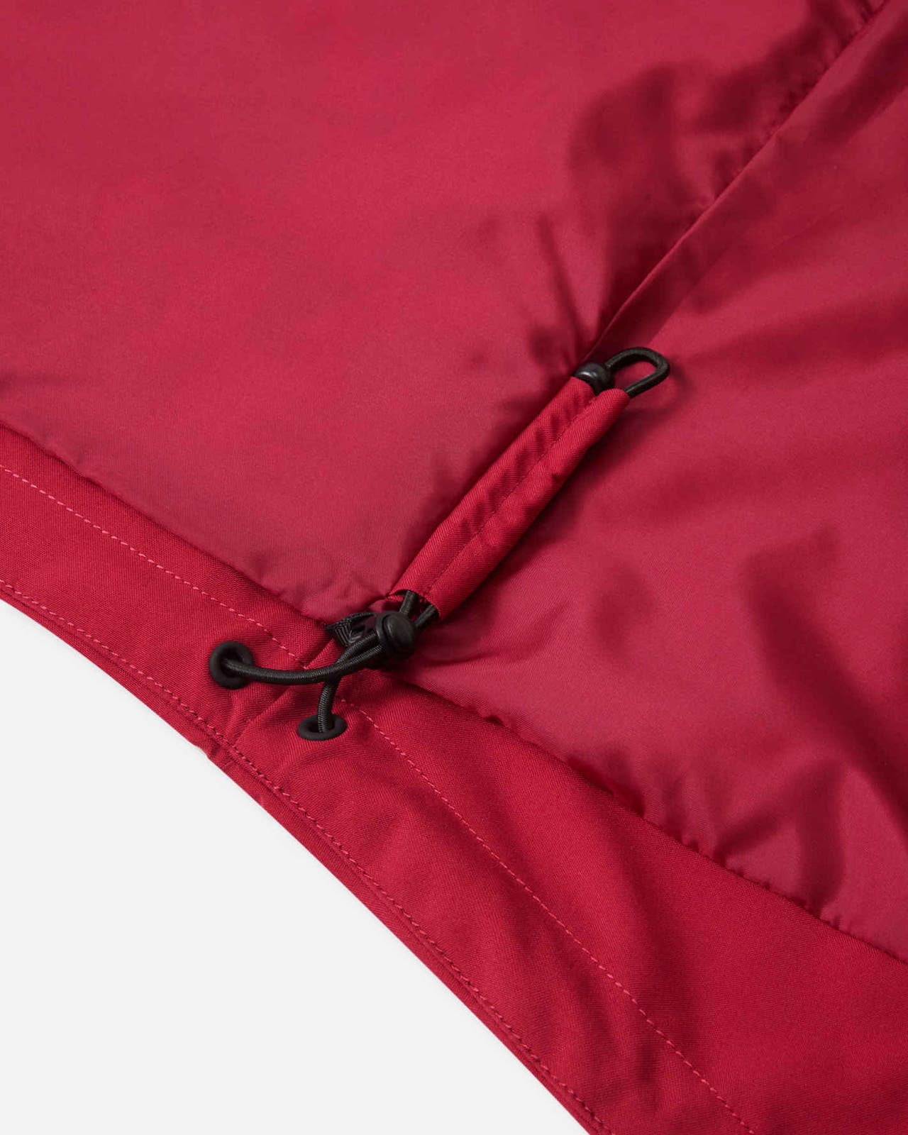 Detailed close-up of the adjustable elastic bungee cord and toggle at the hem of the cranberry red Reima Syddi 3-in-1 kids jacket, designed for a custom fit to seal out cold drafts and snow.