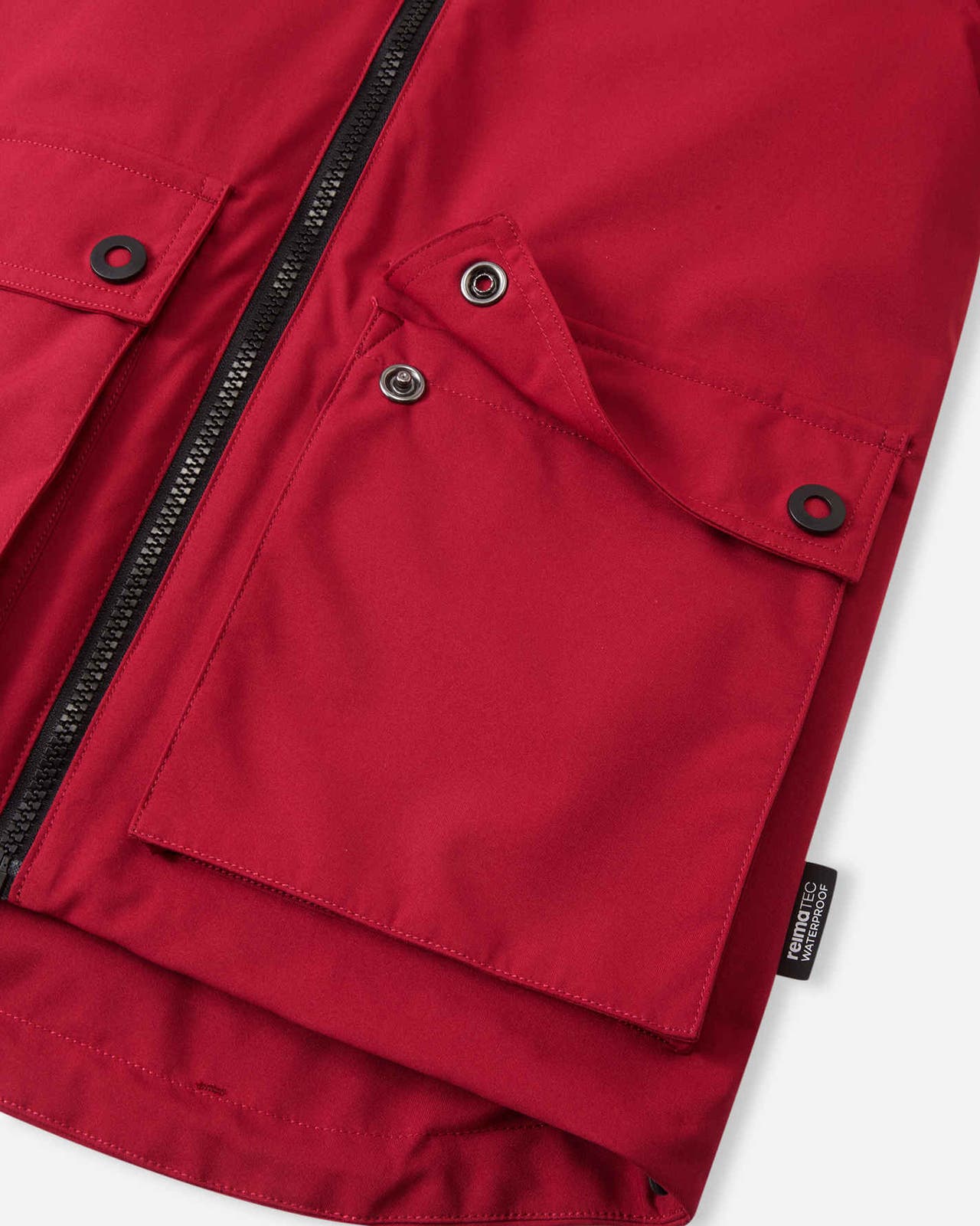 Close-up detail of the cranberry red Reima Syddi 3-in-1 kids jacket, showing the sturdy main front zipper, a spacious snap-flap pocket, and the Reimatec waterproof side tag.