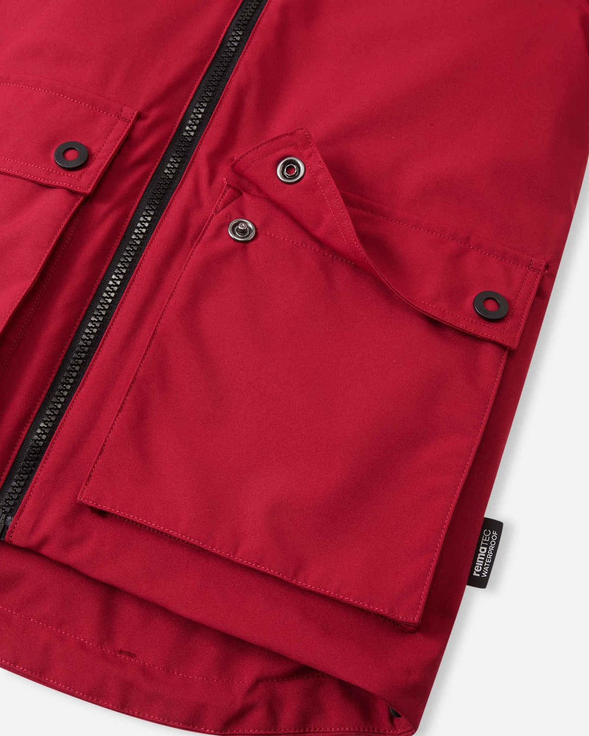 Close-up detail of the cranberry red Reima Syddi 3-in-1 kids jacket, showing the sturdy main front zipper, a spacious snap-flap pocket, and the Reimatec waterproof side tag.
