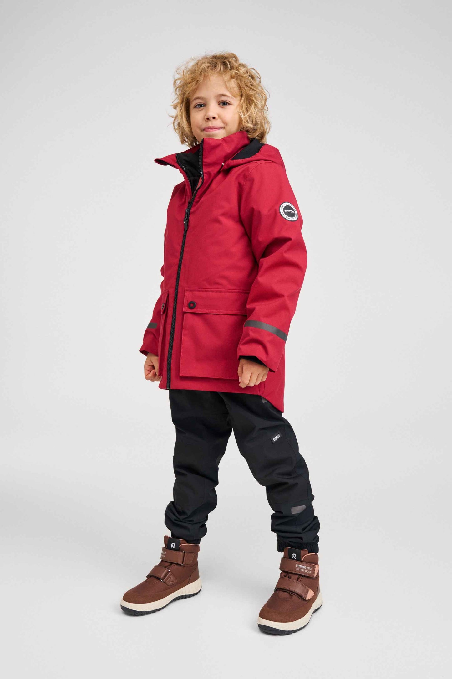 A full-body lifestyle shot of a child modeling the Reima Syddi 3-in-1 kids jacket in cranberry red, paired with black snow pants and winter boots, ready for outdoor winter adventures.
