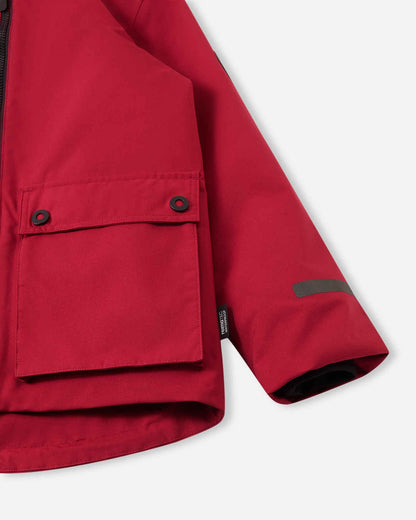 Close-up of the lower half of the cranberry red Reima Syddi 3-in-1 kids jacket, featuring a large buttoned flap pocket, a reflective safety strip on the sleeve, and a waterproof Reimatec brand tag.