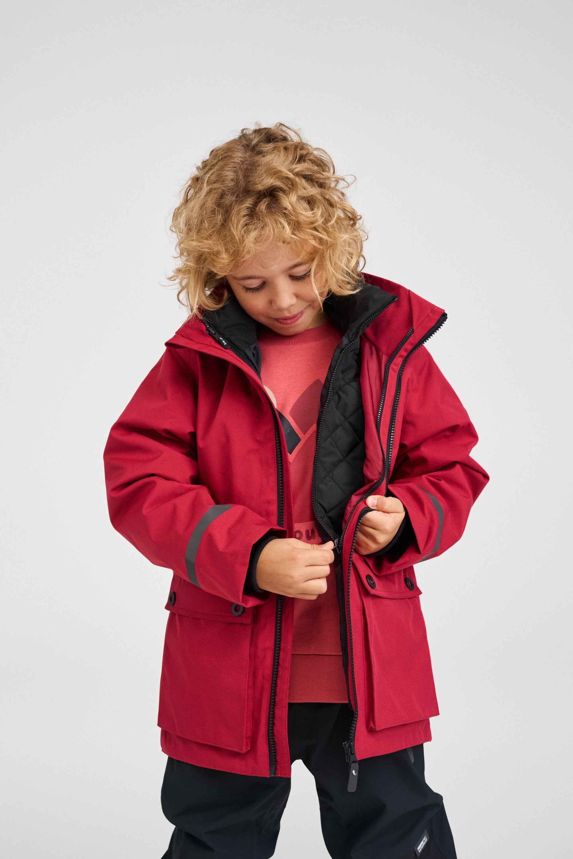 A child unzipping the waterproof cranberry red outer shell of the Reima Syddi 3-in-1 kids jacket to reveal the versatile, detachable black quilted inner jacket underneath.