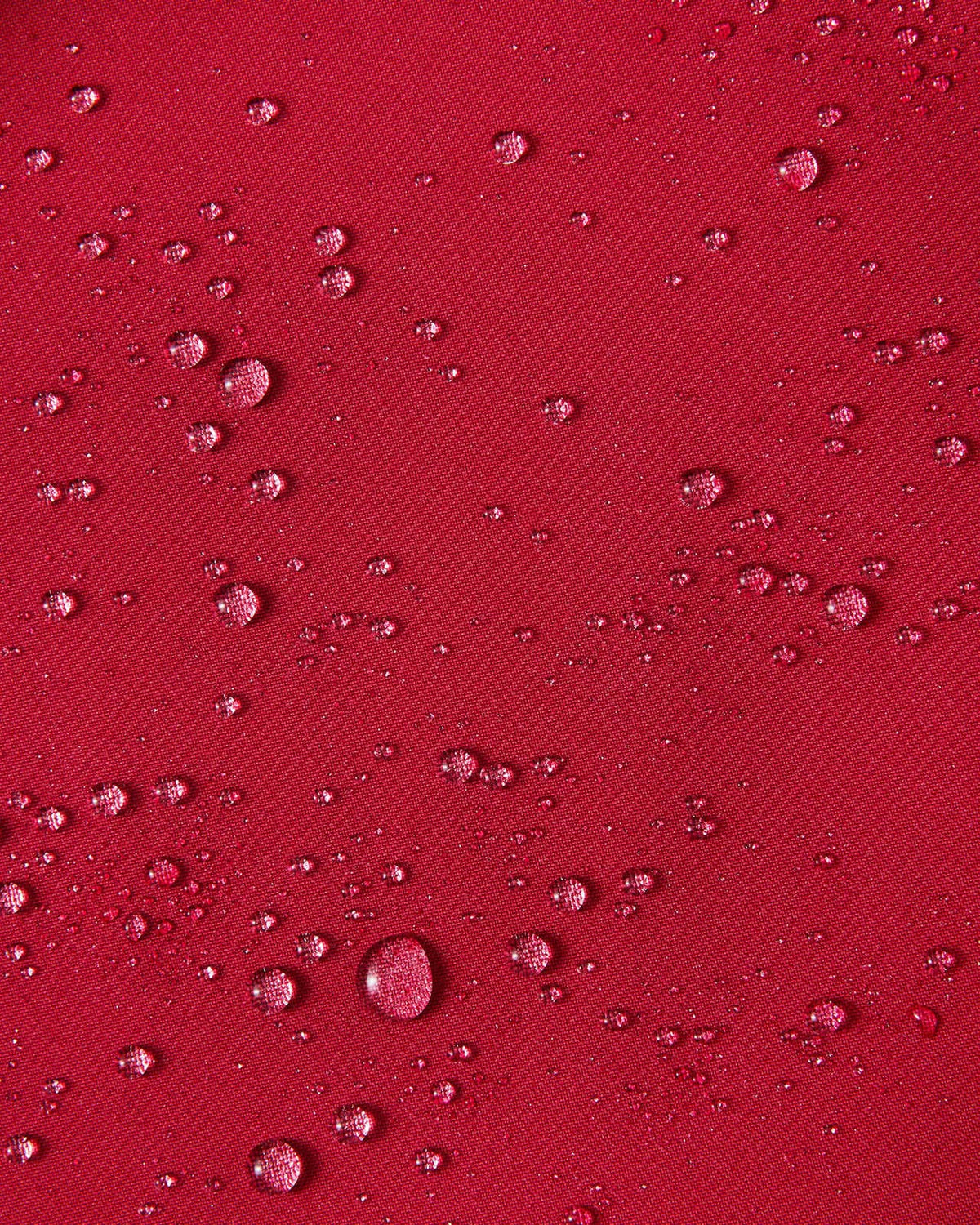 A close-up texture shot demonstrating water droplets cleanly beading up on the cranberry red waterproof exterior fabric of the Reima Syddi 3-in-1 kids jacket.