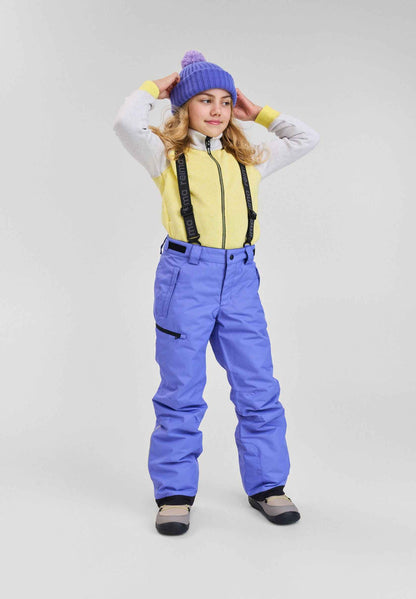 Lifestyle portrait of a young girl wearing the breezy violet Reima Terrie kids' winter ski pants paired with a yellow fleece jacket, purple knit beanie, and snow boots, demonstrating the comfortable fit, mobility, and high visibility on the mountain.