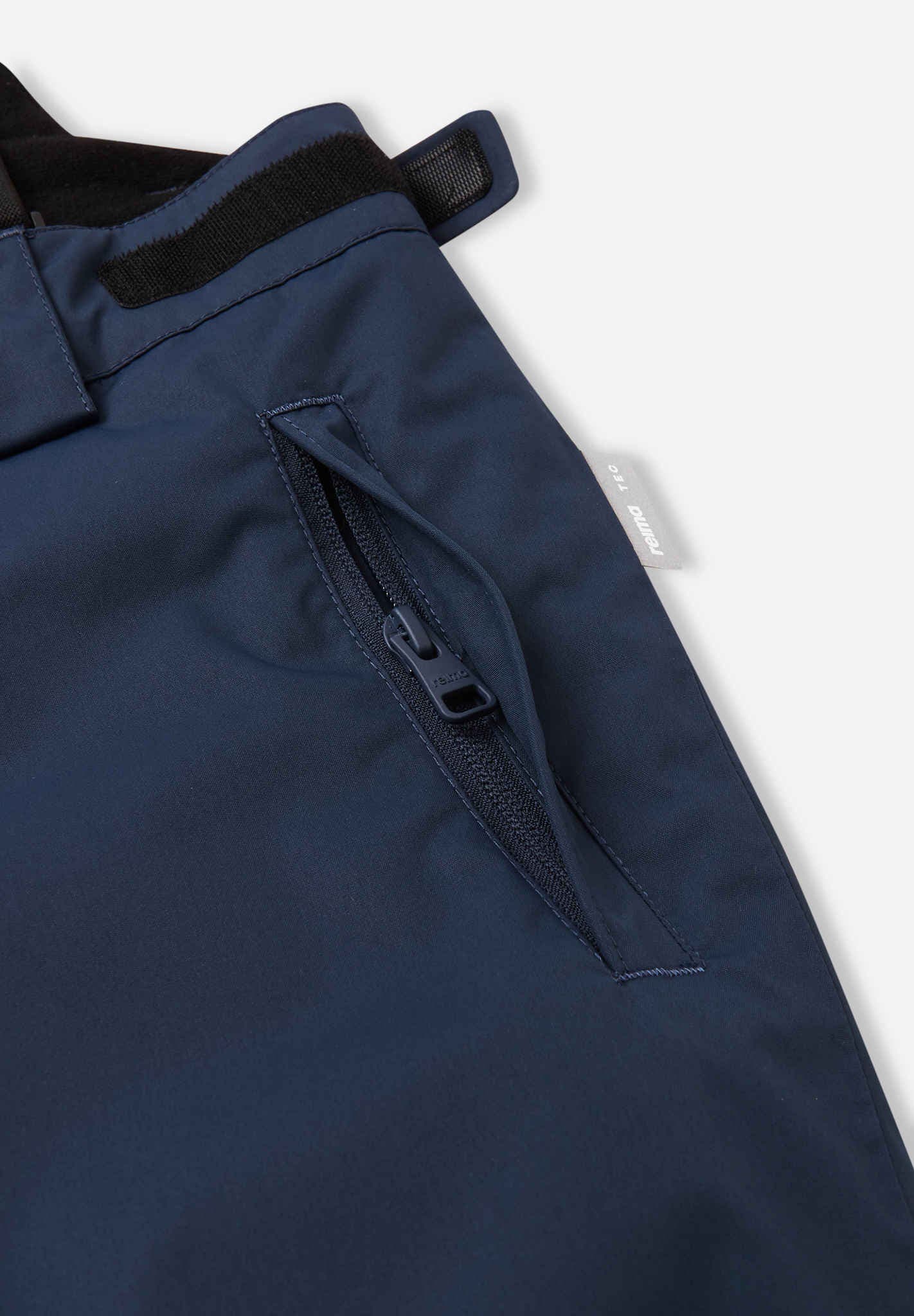 Close-up detail of the hip and thigh on the navy blue Reima Terrie kids' ski pants, highlighting secure zippered pockets and adjustable hook-and-loop waist tabs for a custom, draft-free fit.