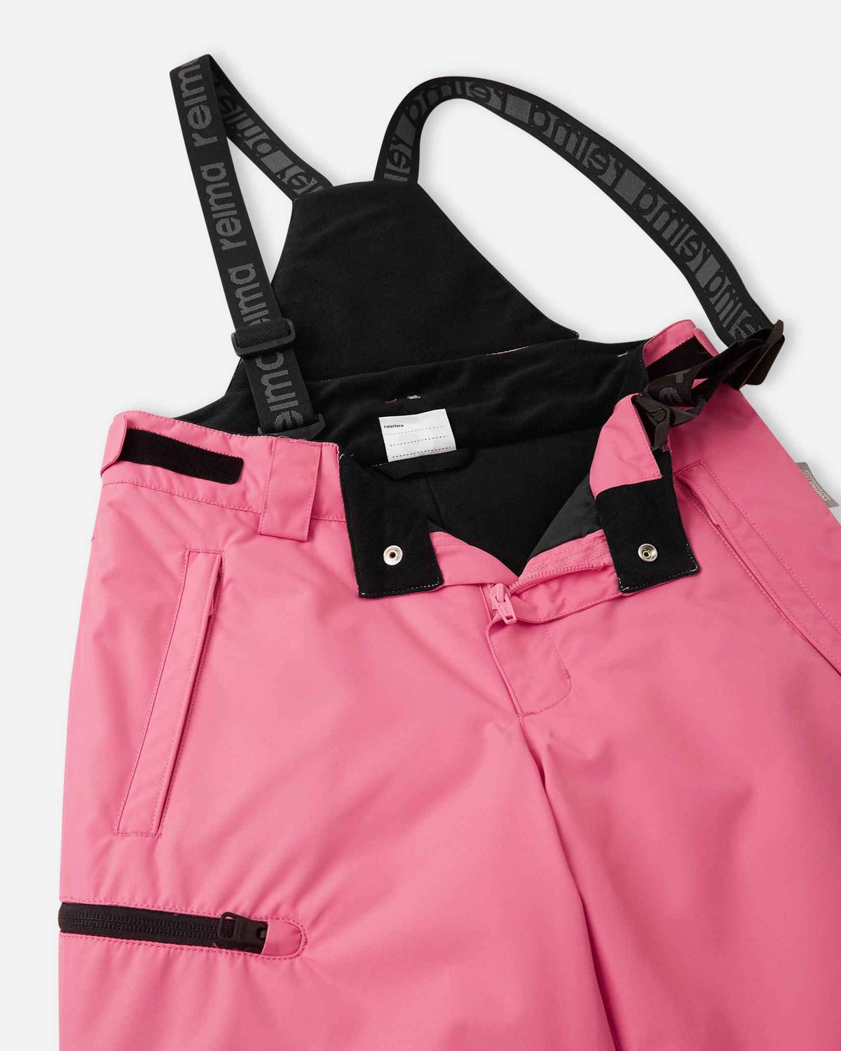 Close-up detail of the front waist on the soft coral Reima Terrie kids' ski pants, showing the secure snap buttons, smooth zipper fly, soft inner lining, and the front attachment points for the adjustable suspenders.