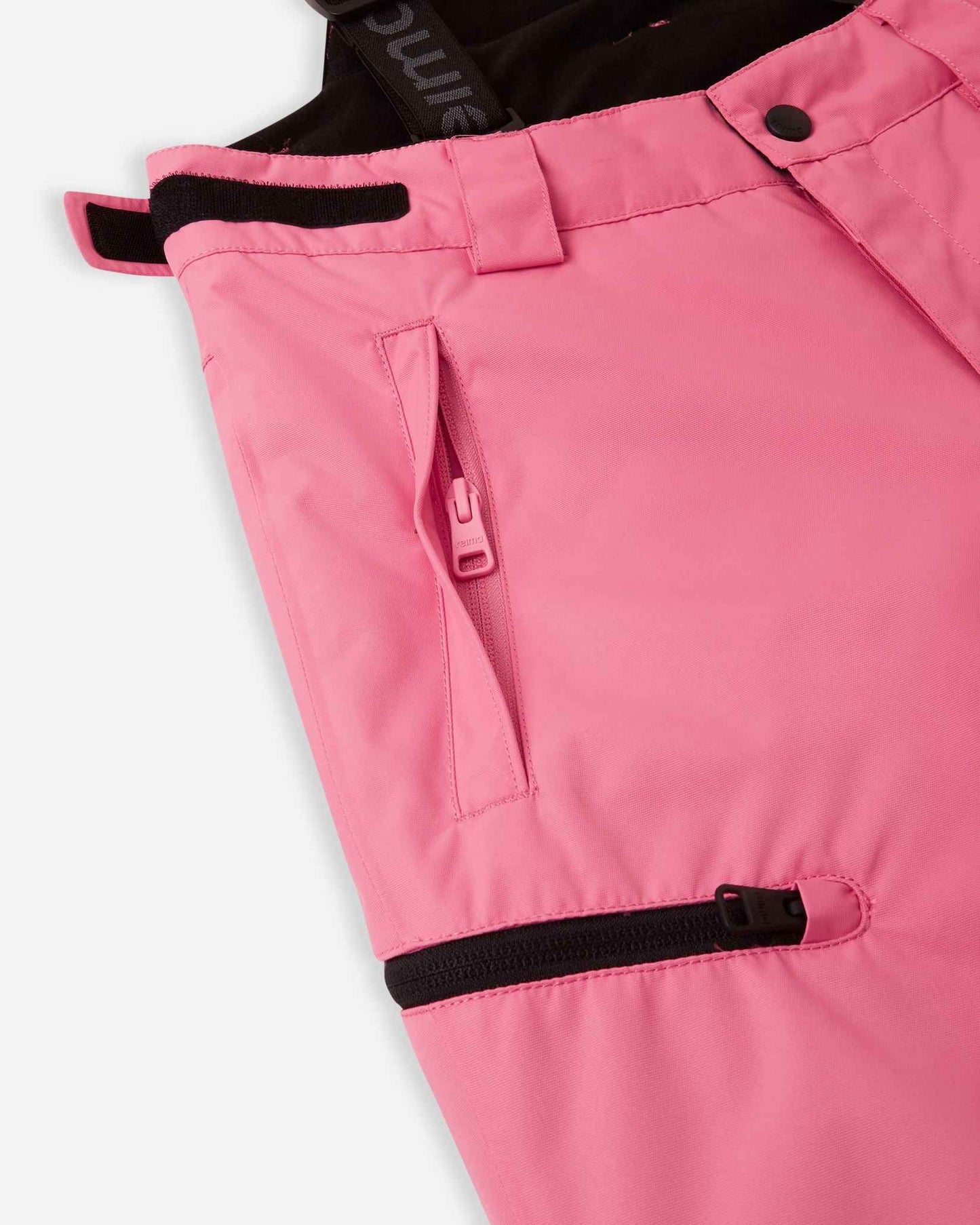 Close-up detail of the hip and thigh on the soft coral Reima Terrie kids' ski pants, highlighting secure zippered pockets and adjustable hook-and-loop waist tabs for a custom, draft-free fit.