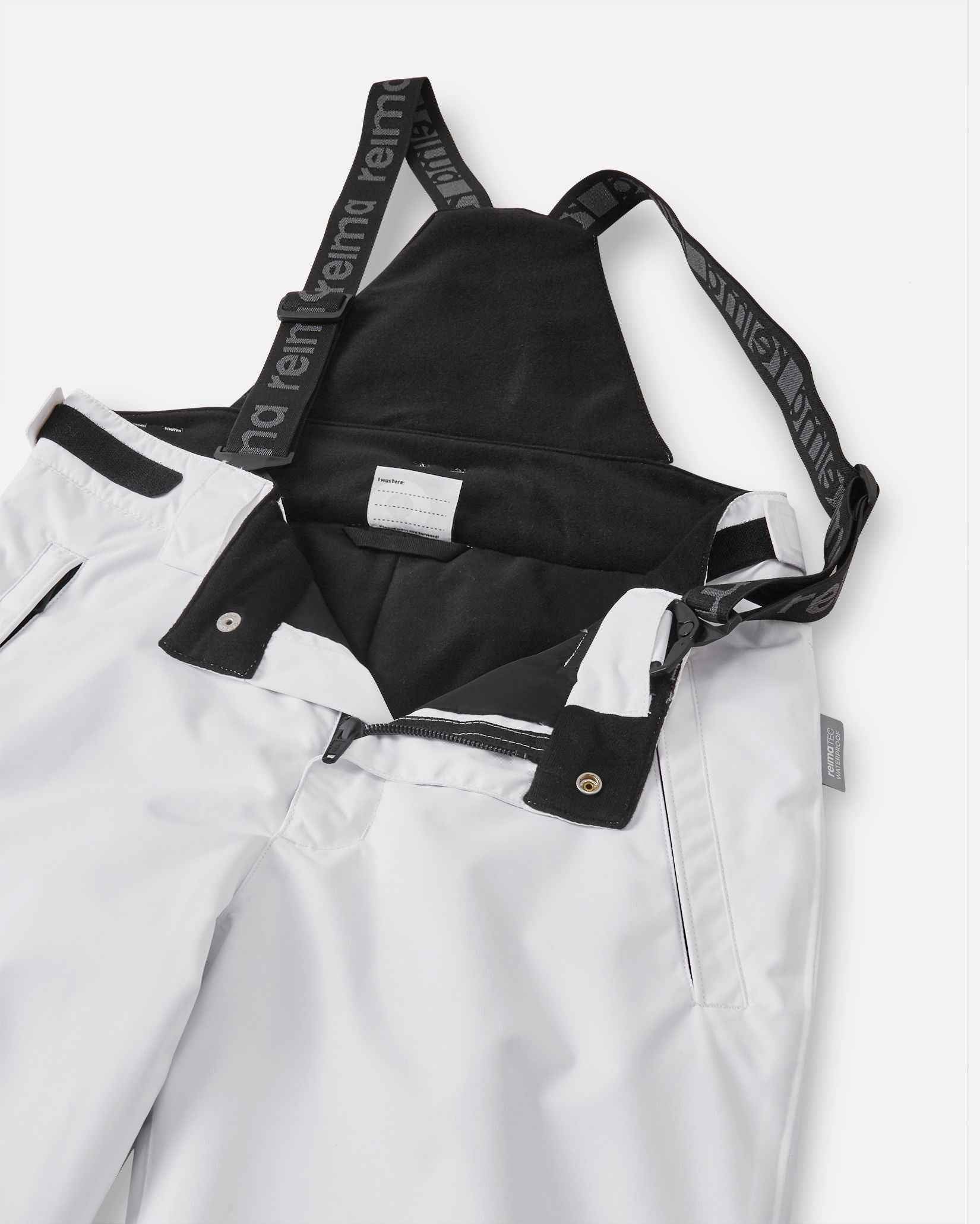 Close-up detail of the front waist on the white Reima Terrie kids' ski pants, showing the secure snap buttons, smooth zipper fly, soft dark inner lining, and the front attachment points for the adjustable suspenders.