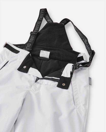 Close-up detail of the front waist on the white Reima Terrie kids' ski pants, showing the secure snap buttons, smooth zipper fly, soft dark inner lining, and the front attachment points for the adjustable suspenders.