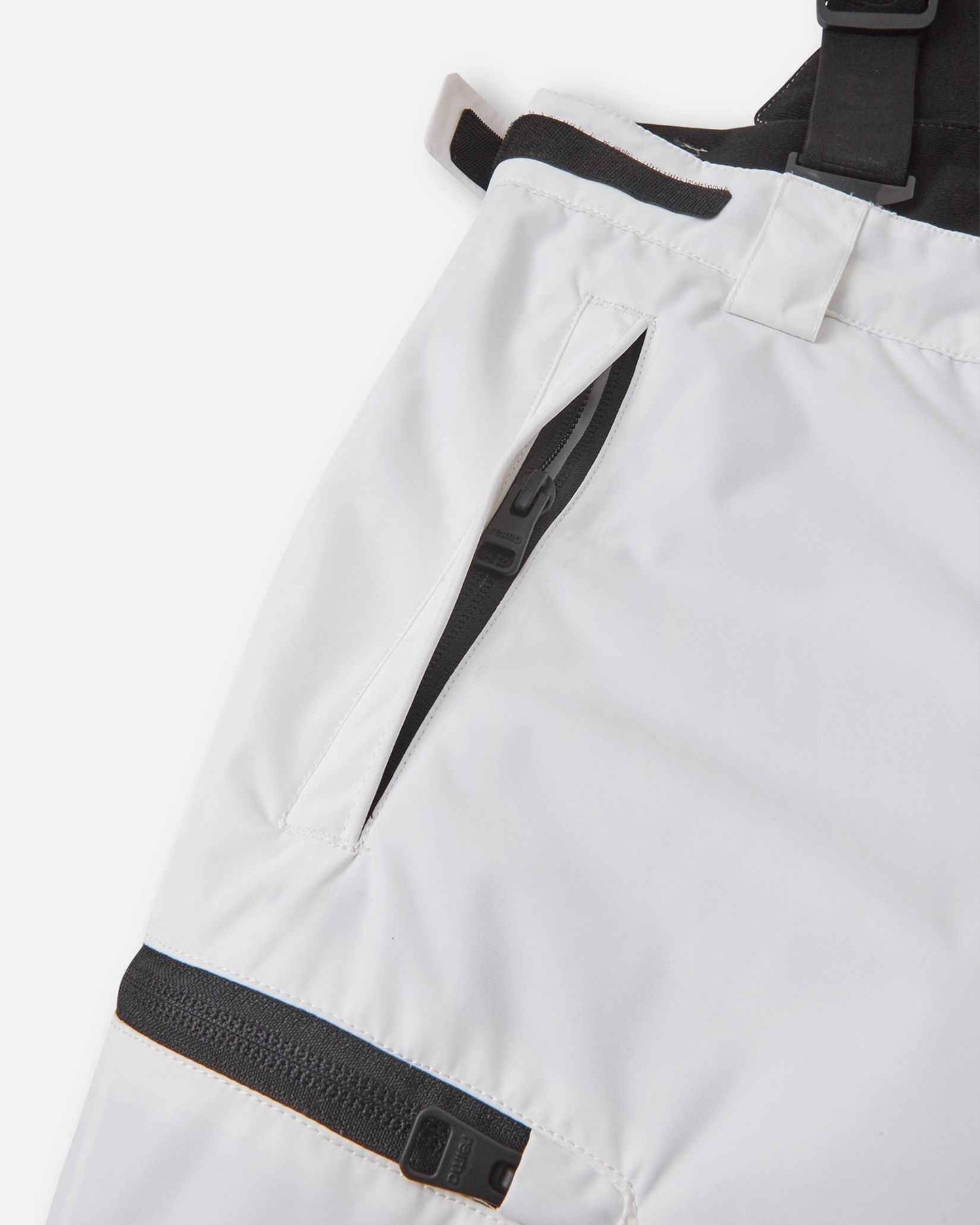 Close-up detail of the hip and thigh on the white Reima Terrie kids' ski pants, highlighting secure black zippered pockets and adjustable hook-and-loop waist tabs for a custom, draft-free fit.