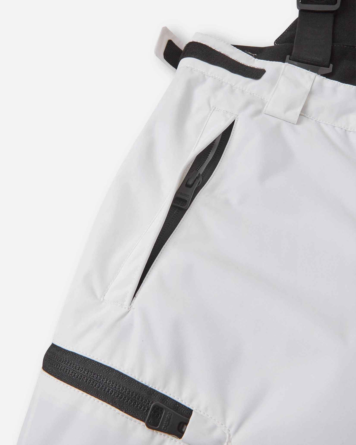 Close-up detail of the hip and thigh on the white Reima Terrie kids' ski pants, highlighting secure black zippered pockets and adjustable hook-and-loop waist tabs for a custom, draft-free fit.