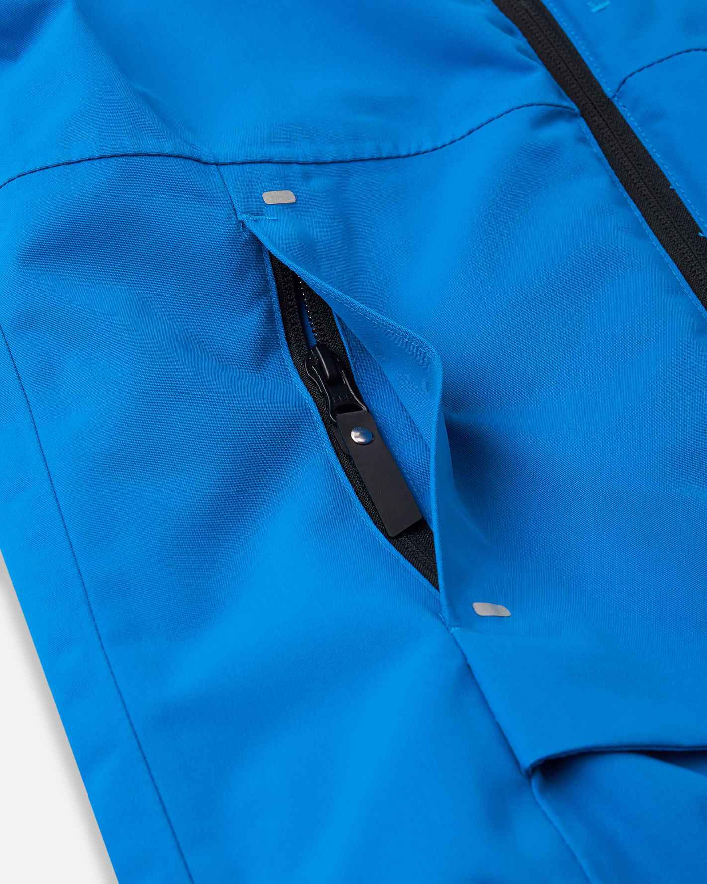 Close-up view of the angled front zippered hand warmer pocket on the bright blue Reima Tieten boys' ski jacket, showing the reinforced zipper flap and reflective bar detailing for secure alpine storage.