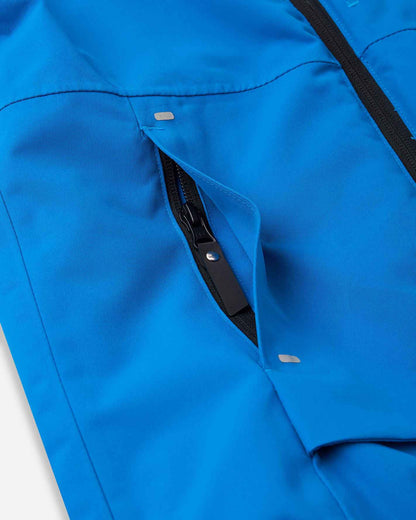 Close-up view of the angled front zippered hand warmer pocket on the bright blue Reima Tieten boys' ski jacket, showing the reinforced zipper flap and reflective bar detailing for secure alpine storage.