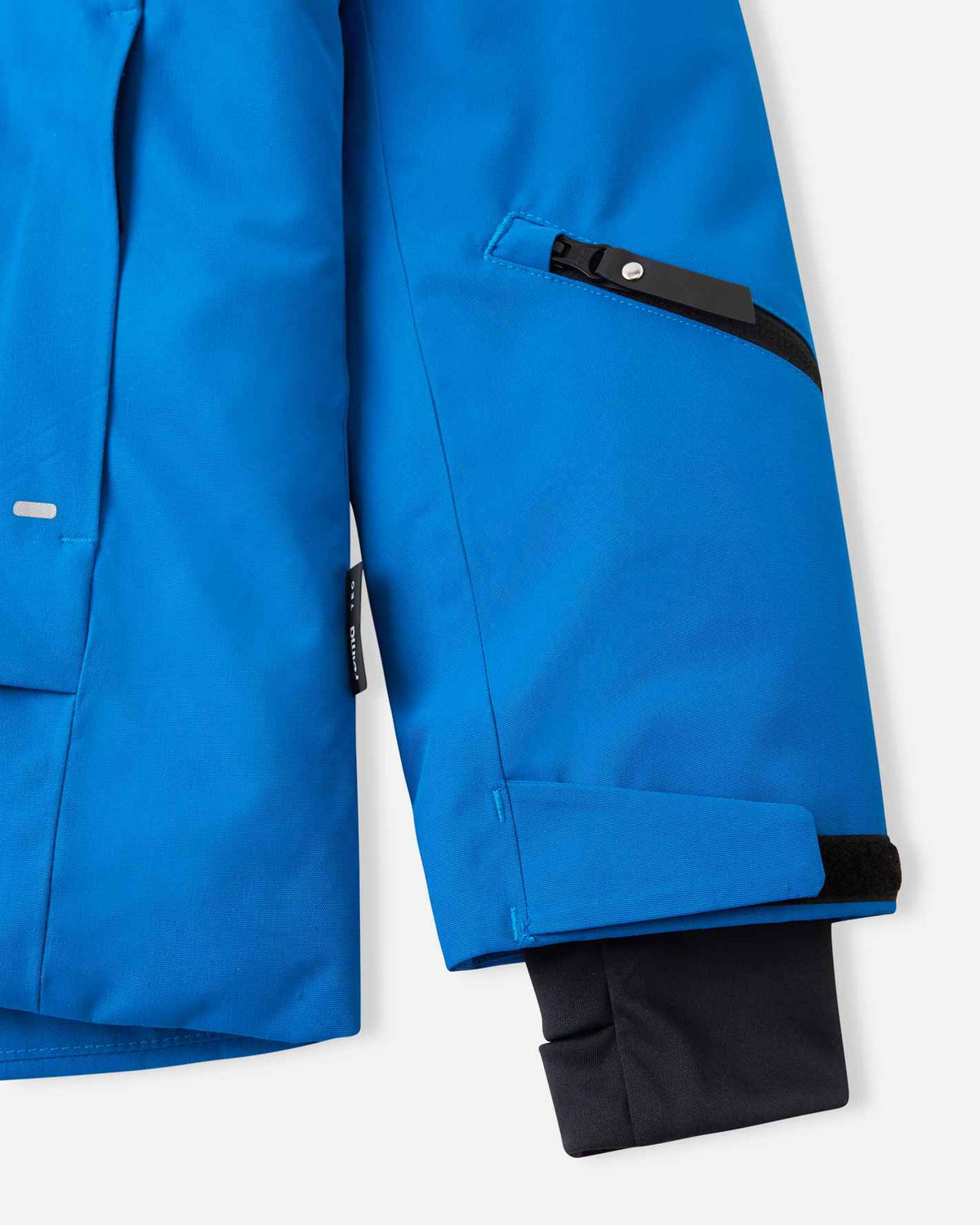 Close-up of the left forearm on the bright blue Reima Tieten boys' snow jacket, featuring a black zippered lift pass pocket, adjustable wrist strap, and an inner thumbhole lycra cuff to seal out snow.