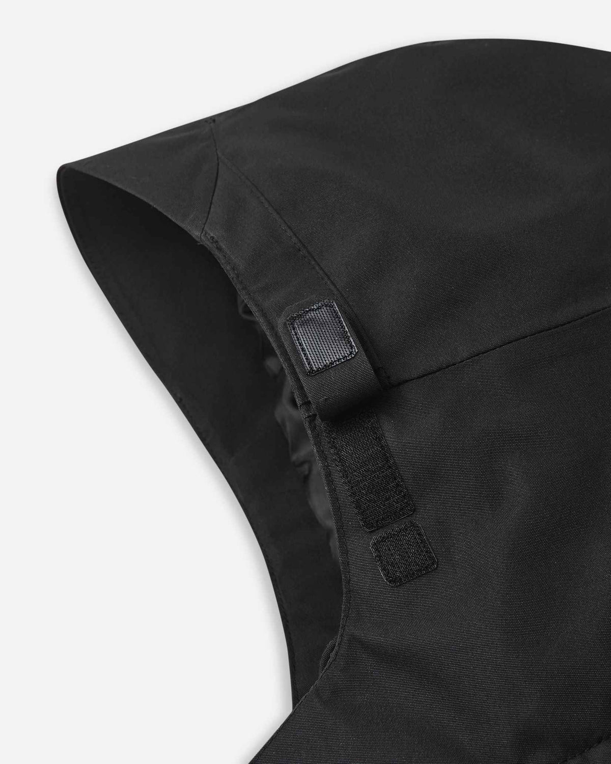 Macro view of the front edge of the hood on the black Reima Tieten youth snow jacket, focusing on the reinforced hook-and-loop tabs that secure the hood firmly to the collar during active downhill runs.