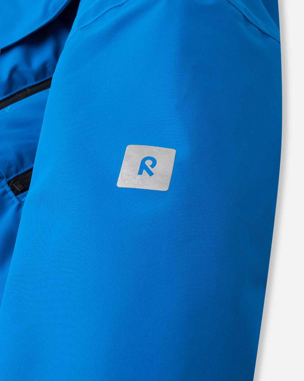 Macro detail of the reflective Reima 'R' safety logo on the bright blue sleeve of the Reima Tieten youth winter coat, designed to enhance low-light visibility during evening ski runs and outdoor play.