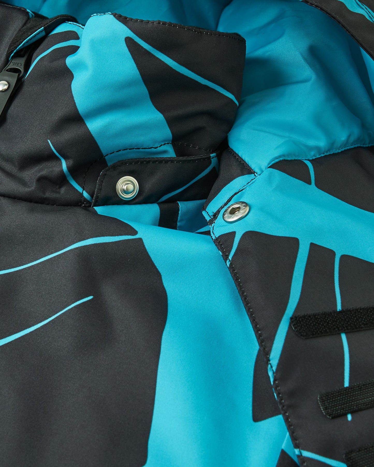 Detailed view of the protective chin guard, storm flap, and secure collar snap buttons on the black Reima Tirro kids' waterproof ski jacket, keeping wind and snow out.