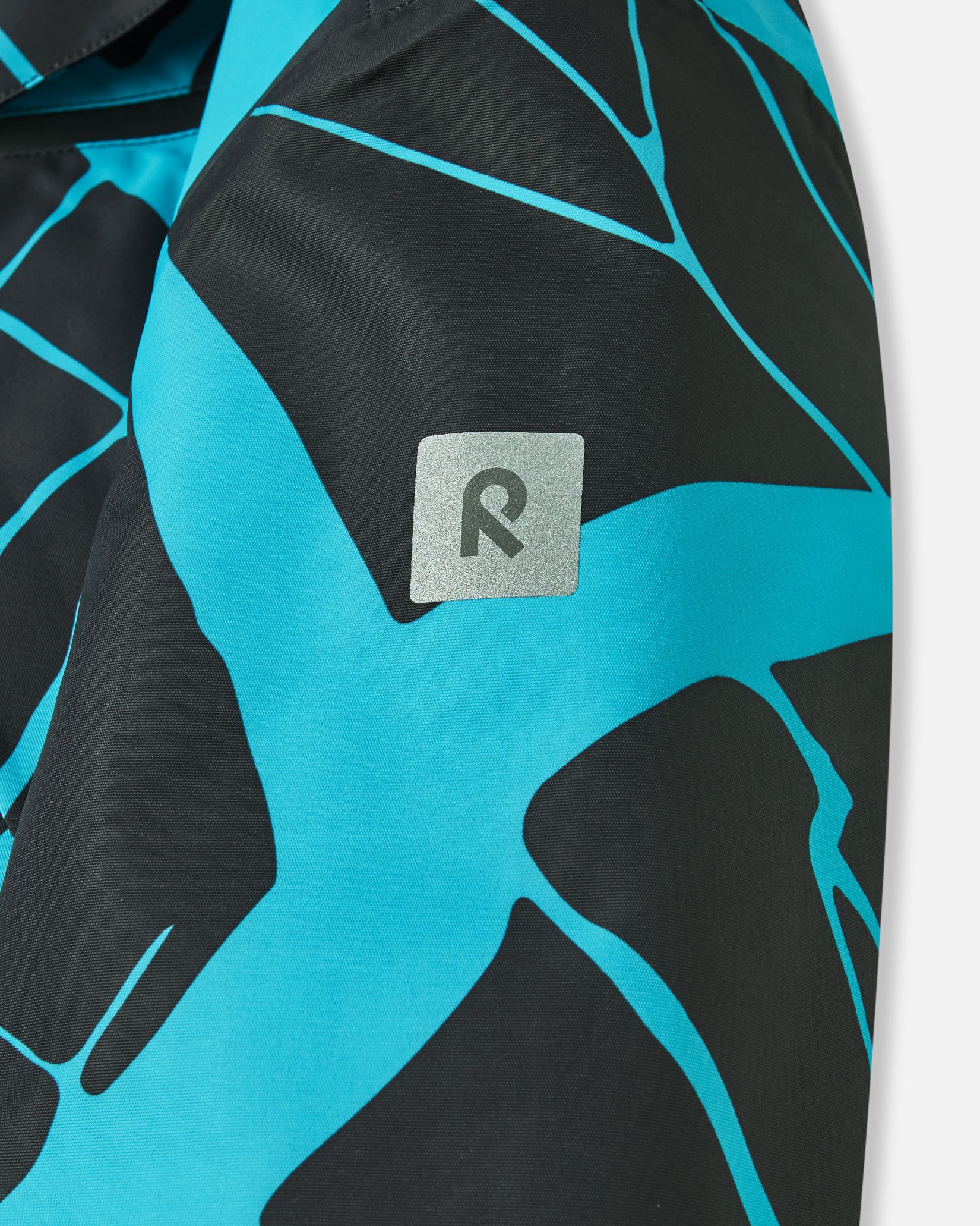 Close-up of the safety-enhancing reflective Reima logo patch on the sleeve of the black Tirro kids' waterproof ski jacket, providing visibility in low-light winter conditions.