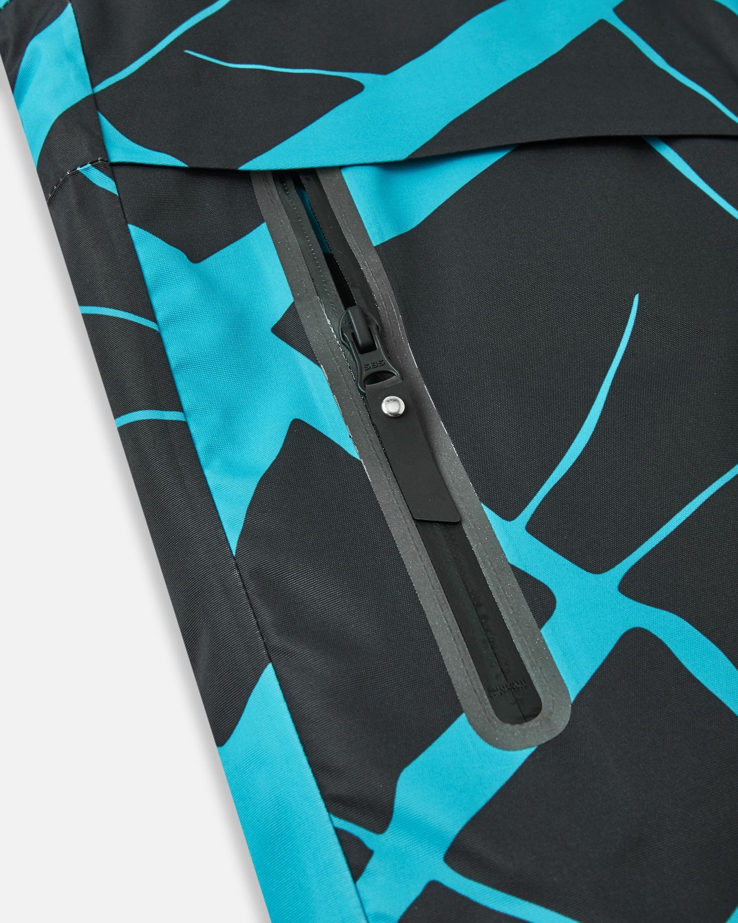 Close-up of the sealed, waterproof zippered side pocket on the black Reima Tirro kids' ski jacket, highlighting the durable, weather-resistant construction and dynamic blue geometric pattern.