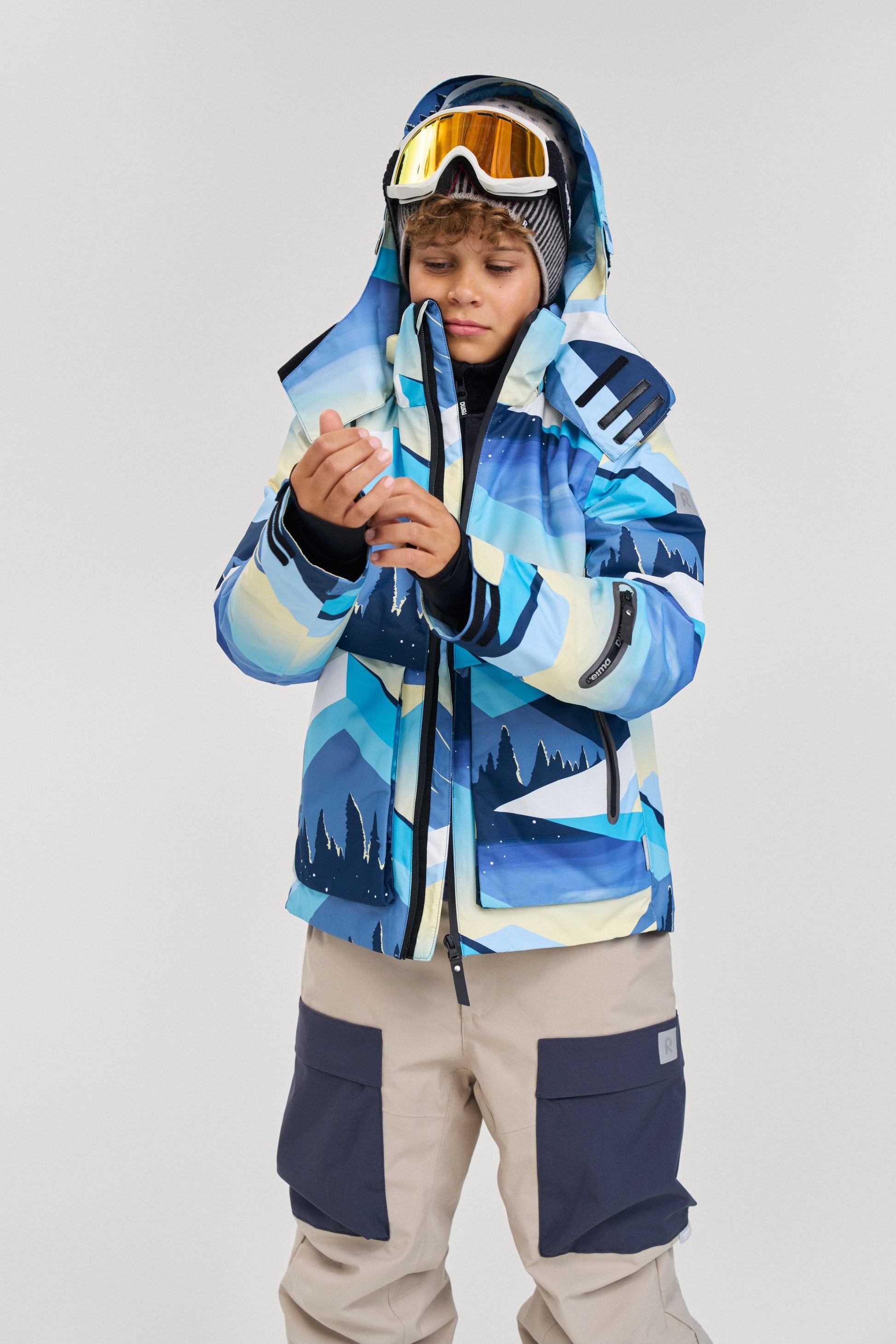Lifestyle portrait of a young boy wearing the ocean blue Reima Tirro kids' waterproof ski jacket, featuring an active mountain pattern and a helmet-compatible adjustable hood for winter sports.
