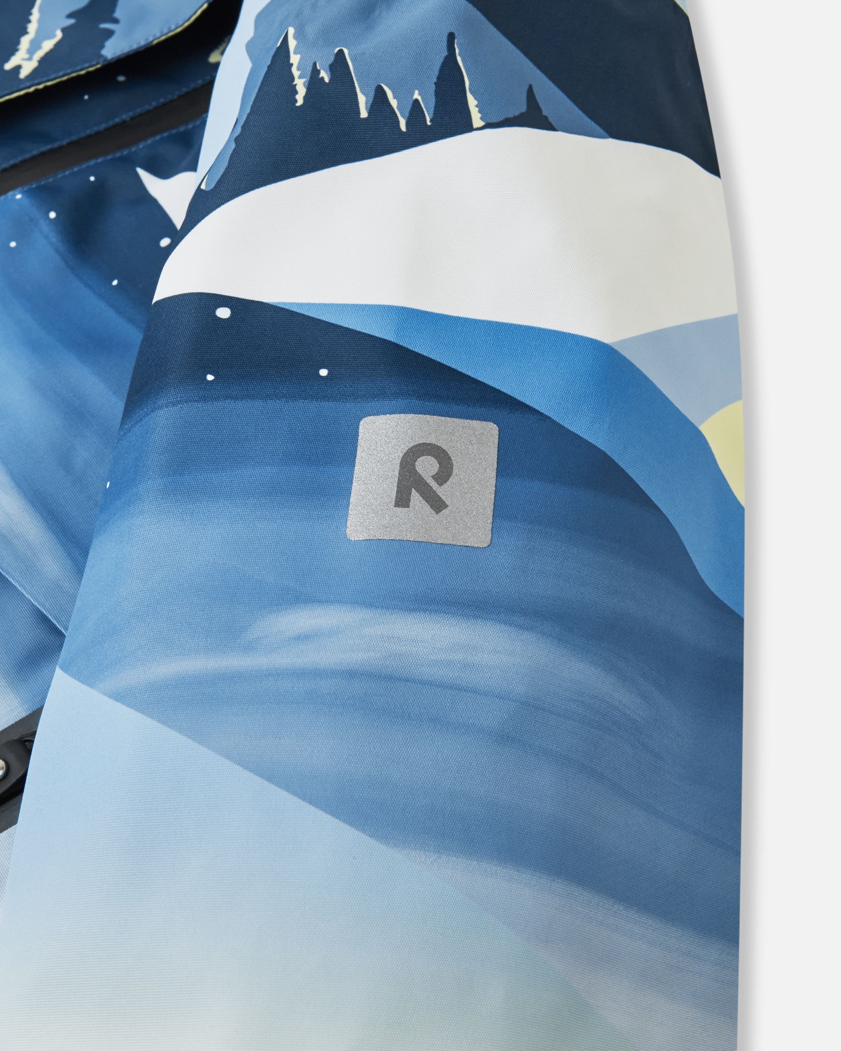 Close-up of the safety-enhancing reflective Reima logo on the sleeve of the ocean blue Tirro kids' waterproof ski jacket.
