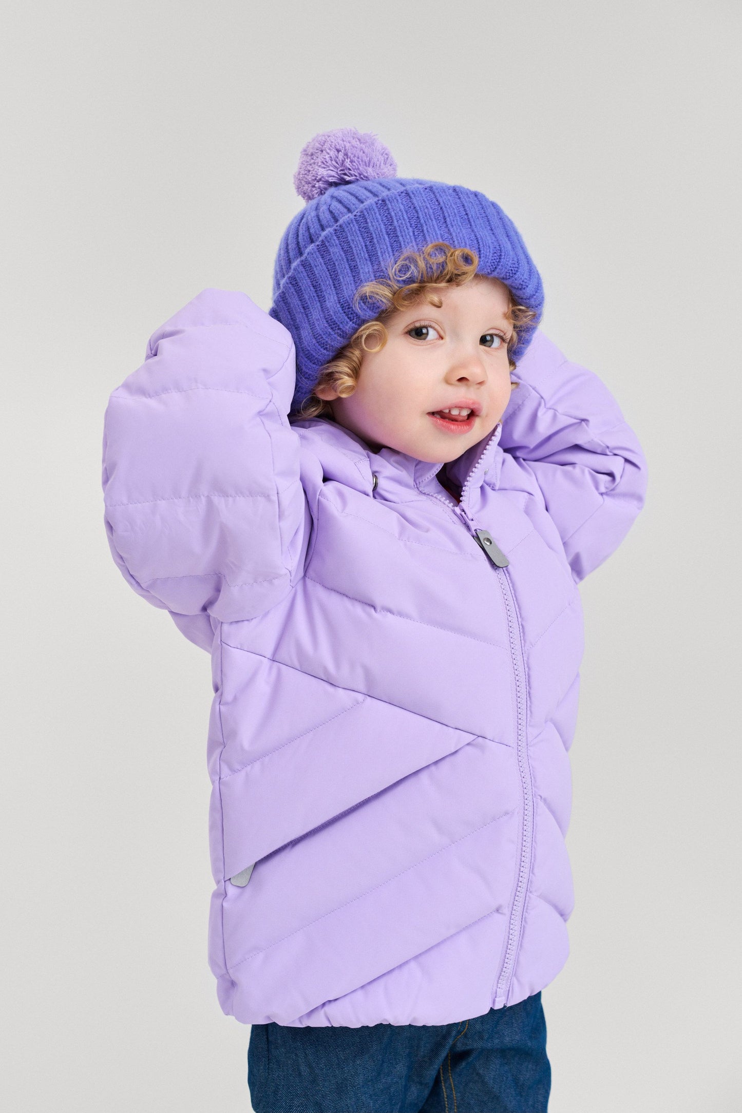 Lifestyle portrait of a young toddler looking over her shoulder while wearing the breezy violet Reima Topsu kids' wool blend beanie paired with a light purple puffer jacket. The image demonstrates how the wide brim provides a secure, warm fit around the ears for active winter play.