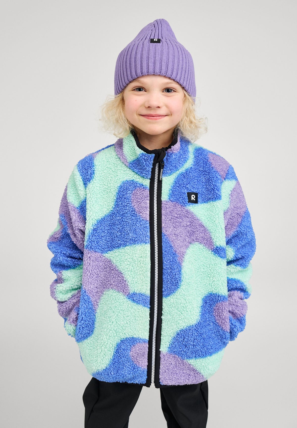 Lifestyle shot of a smiling child wearing the sparkly blue Reima Tupsis teddy fleece jacket paired with a purple beanie, demonstrating the jacket's comfortable fit and stylish urban look.