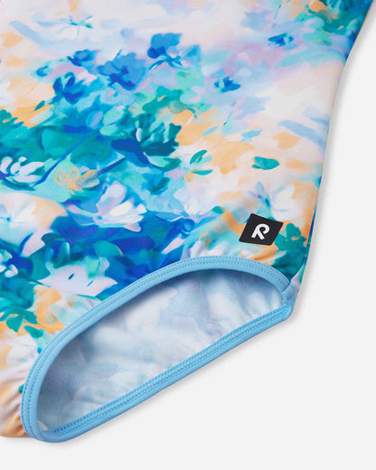 Detailed close-up of the Reima Uimaan girls' cool blue watercolor swimsuit, focusing on the soft, quick-drying stretch fabric, light blue elastic leg binding, and the signature Reima logo tag.