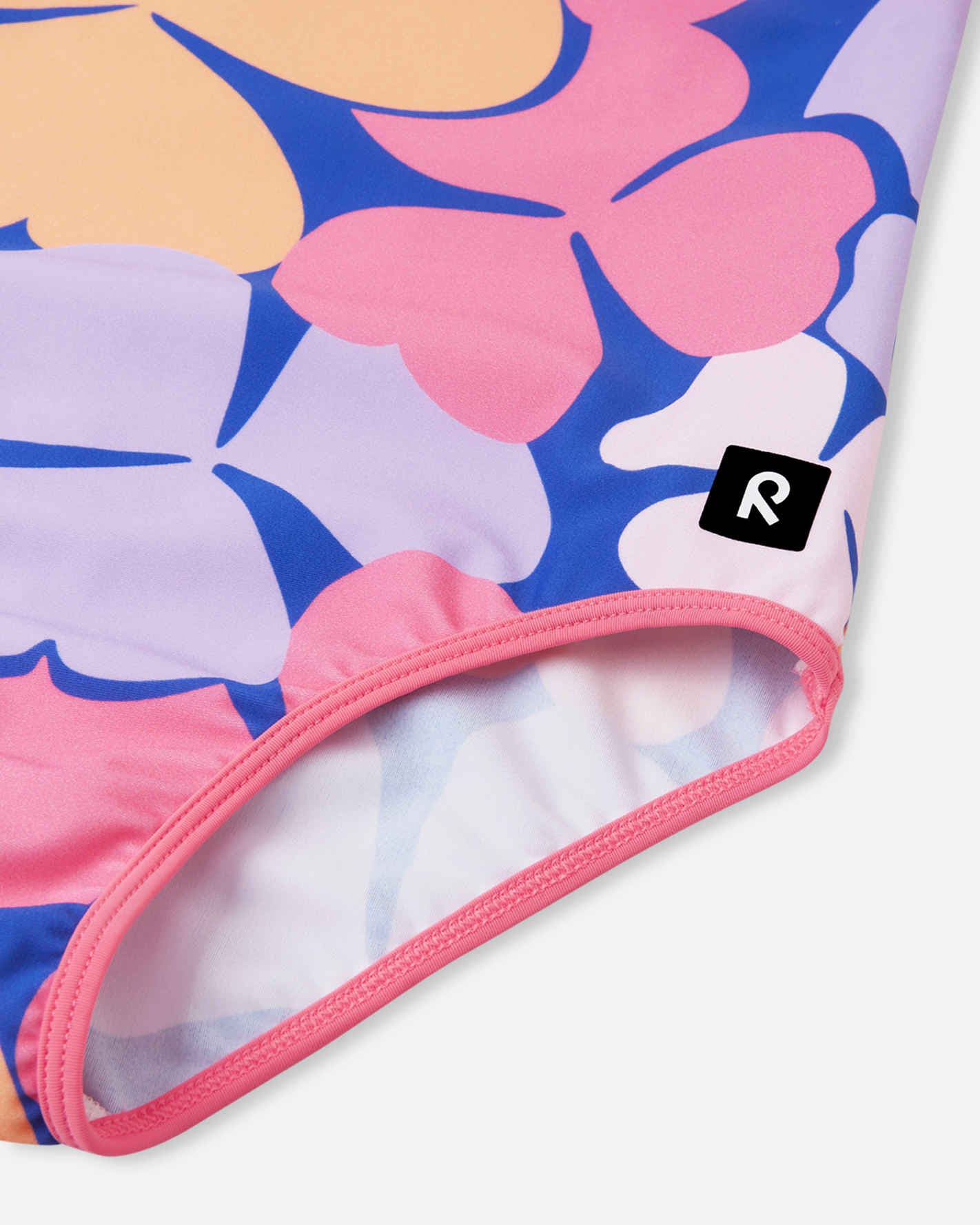 Close-up detail of the Reima Uimaan girls' soft coral floral swimsuit, highlighting the smooth, stretchy leg opening with pink contrast binding and the small black Reima logo patch.
