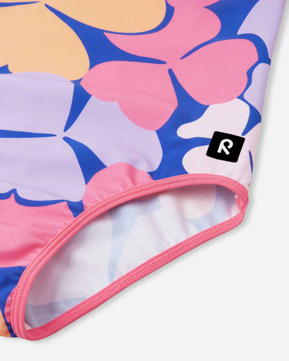 Close-up detail of the Reima Uimaan girls' soft coral floral swimsuit, highlighting the smooth, stretchy leg opening with pink contrast binding and the small black Reima logo patch.