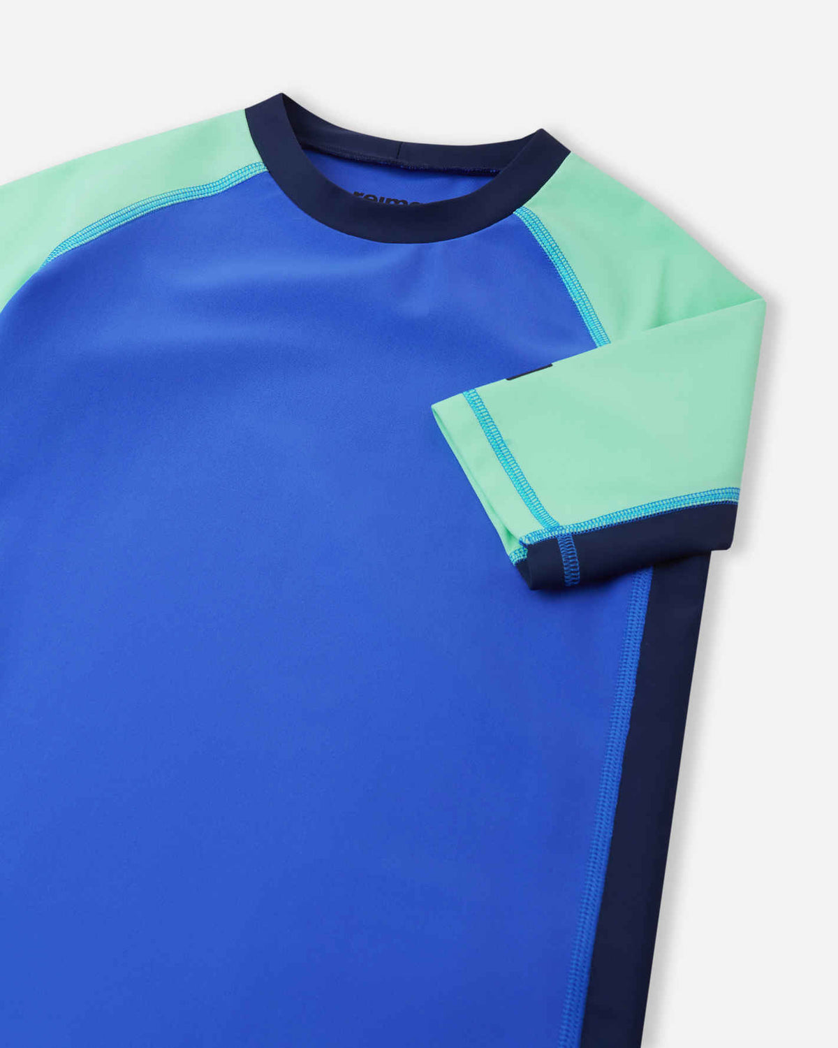 Close-up detail of the Reima Uiva kids' sunproof rash guard in sparkly blue, highlighting the stretchy mint green raglan sleeve, durable flatlock seams, and a protective high crew neckline.