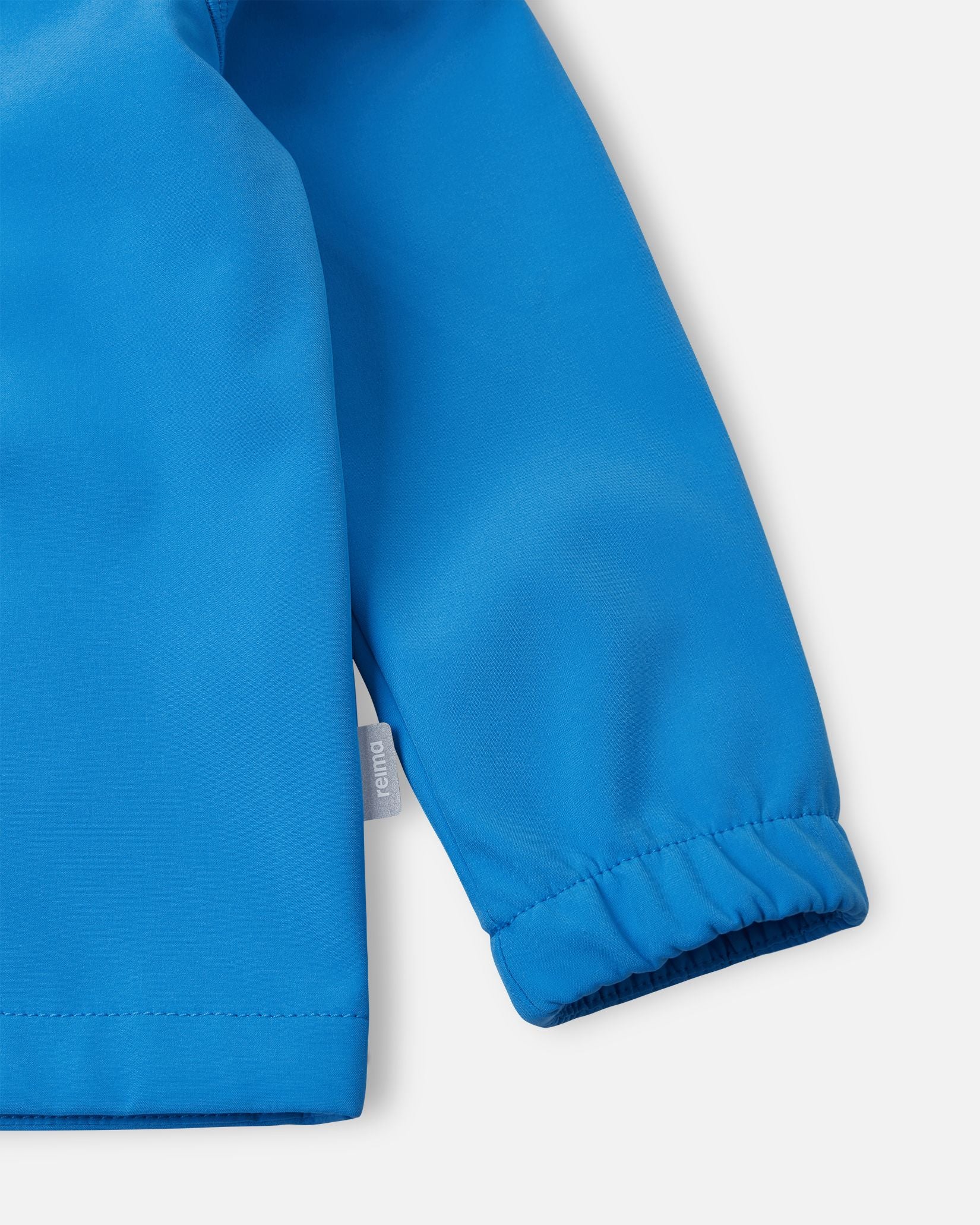Detailed close-up of the sleeve on the cool blue Reima Vantti kids' softshell jacket, highlighting the durable windproof fabric, the small Reima logo side tag, and the snug elastic cuff that seals out the elements.