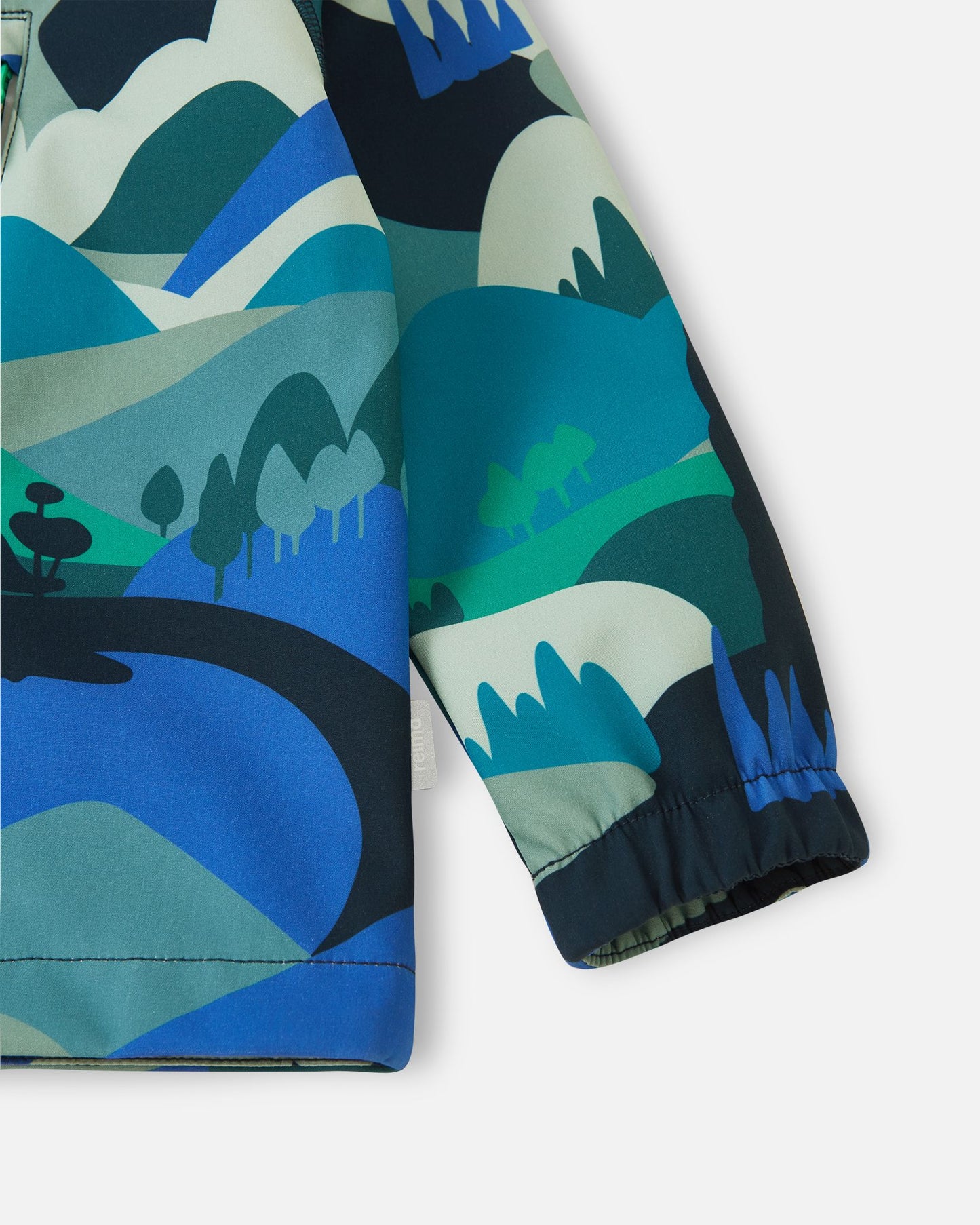 Detailed close-up of the lower body and sleeve on the sparkly blue Reima Vantti kids' softshell jacket, focusing on the vibrant mountain print, side logo tag, and snug wind-blocking elastic cuff.