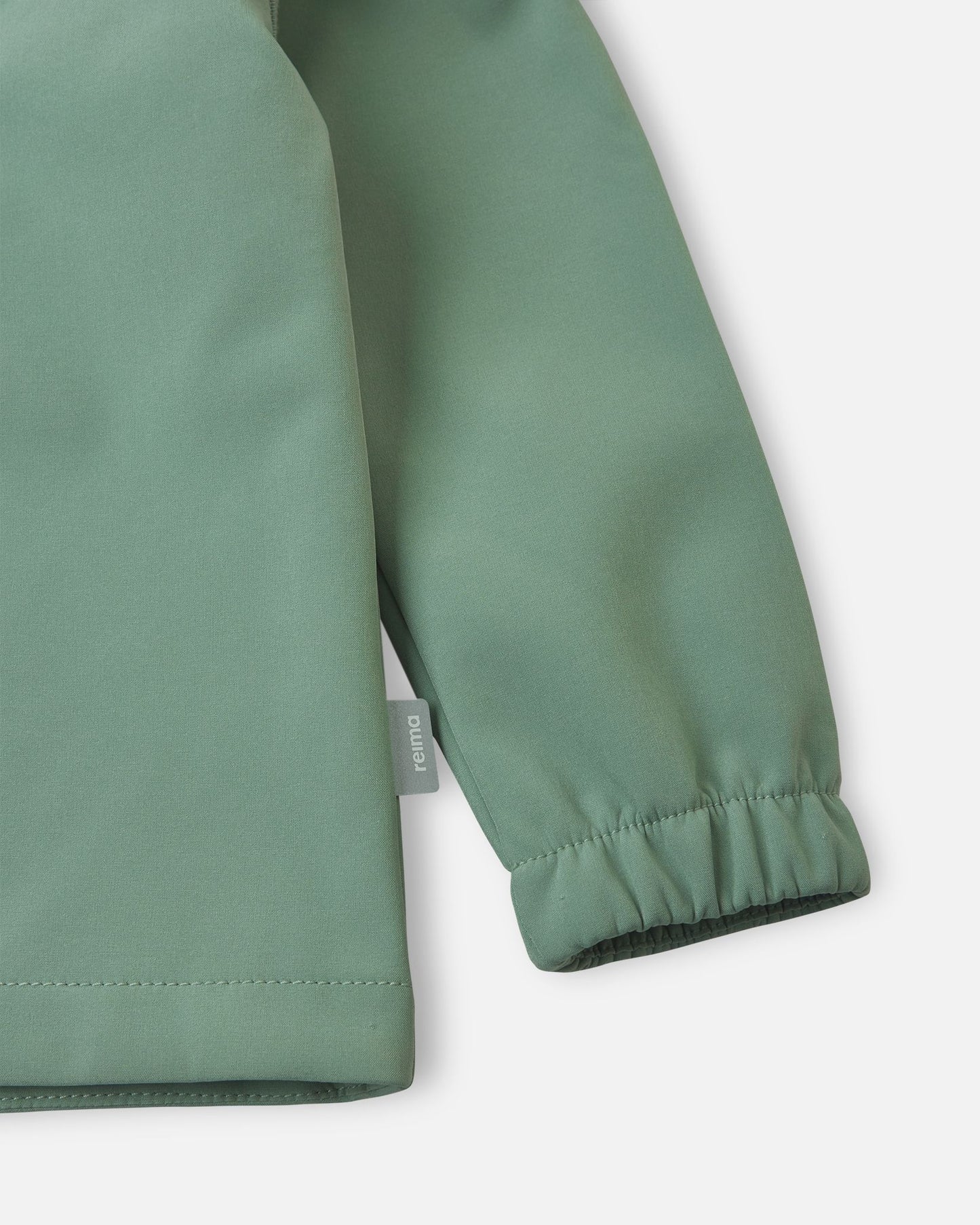 Detailed close-up of the sleeve on the stone green Reima Vantti kids' softshell jacket, highlighting the durable windproof fabric, the small Reima logo tag, and the snug elastic cuff.