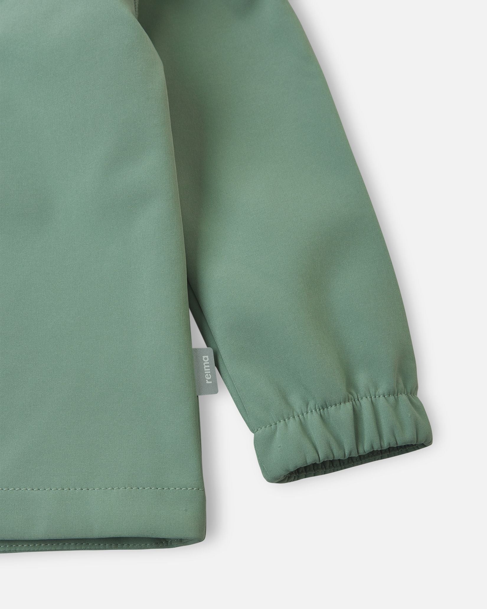 Detailed close-up of the sleeve on the stone green Reima Vantti kids' softshell jacket, highlighting the durable windproof fabric, the small Reima logo tag, and the snug elastic cuff.