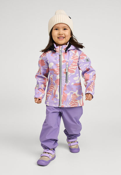Lifestyle shot of a smiling young girl wearing the blooming lilac Reima Vantti kids' softshell jacket paired with matching purple outdoor pants and a cream beanie, demonstrating its comfortable and stylish fit for everyday outdoor play.