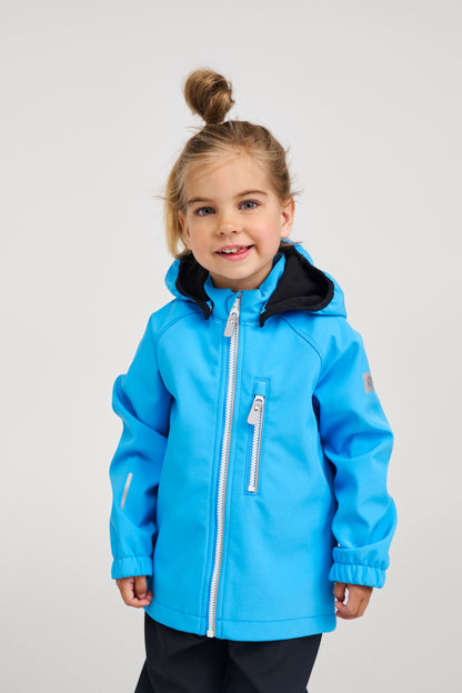 Lifestyle shot of a smiling young girl wearing the cool blue Reima Vantti kids' softshell jacket, demonstrating its comfortable, flexible fit and stylish design for everyday outdoor play and school wear.