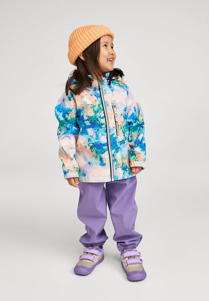 Lifestyle shot of a smiling young girl wearing the fresh blue floral Reima Vantti kids' softshell jacket paired with purple outdoor pants and an orange beanie, demonstrating its comfortable and stylish fit for everyday play.