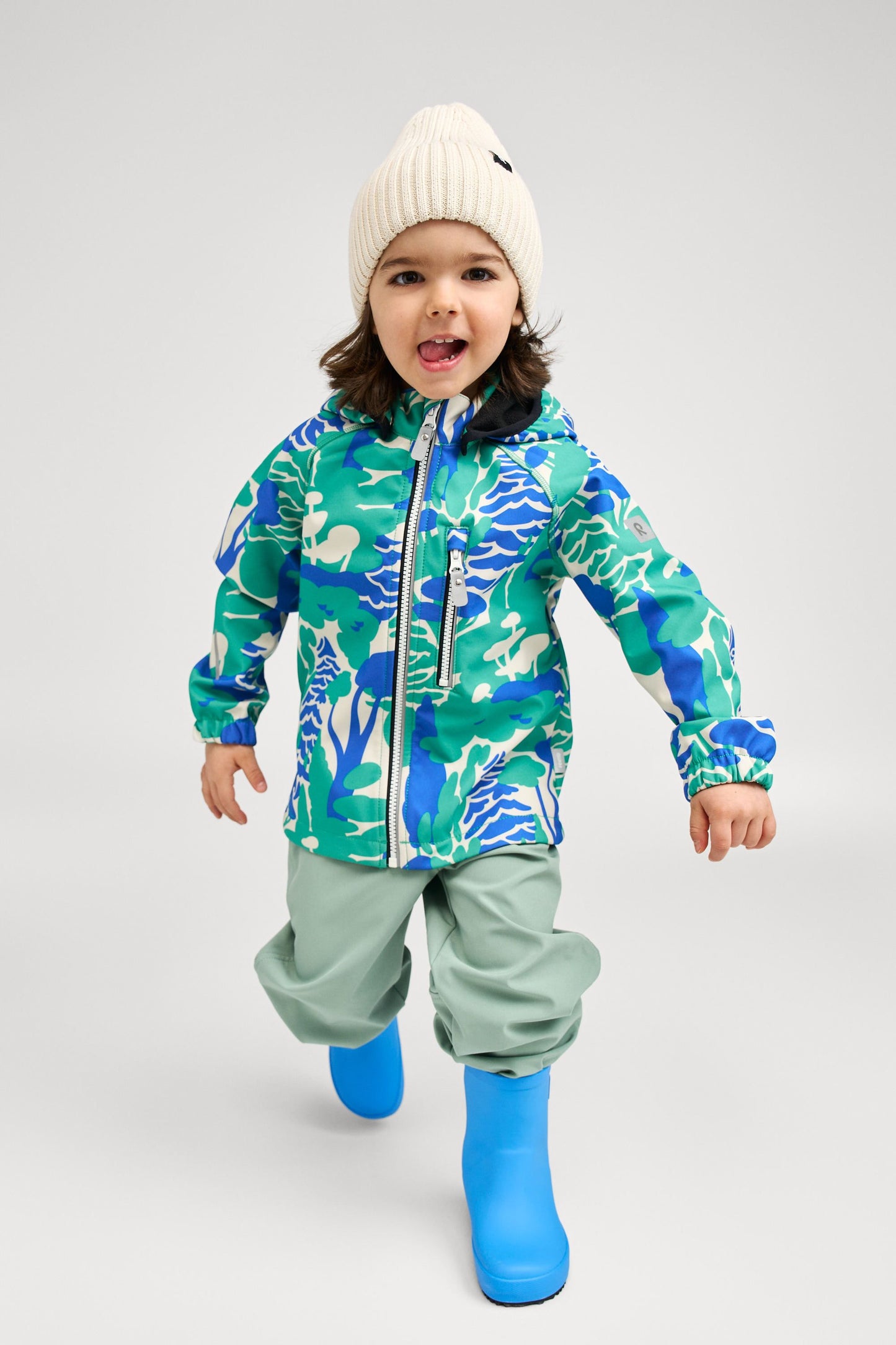 Lifestyle shot of an active toddler wearing the green forest print Reima Vantti kids' softshell jacket paired with light green rain pants and blue boots, demonstrating its comfortable and protective fit for rainy day play.